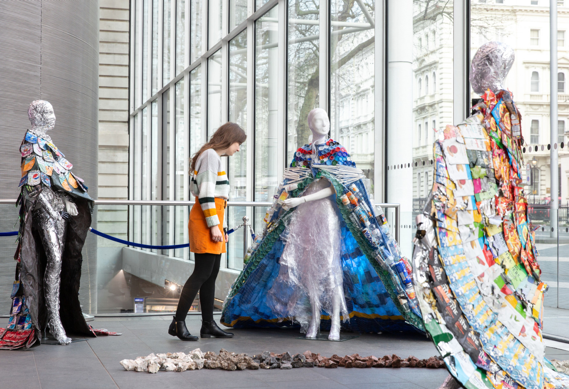 Exhibition shows three manequins wearing long trailing cloaks made from street litter. One made from plastic drinks bottles and aluminium cans, with a dress wrap of cling film, another made from colourful crisp packets. A young white woman with long brown wavy hair wearing an orange skirt and jumper  is looking closely at one.