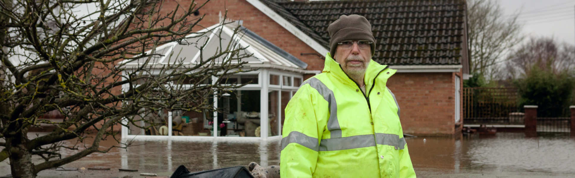 man in high viz standing thigh deep in flooded surroundings with house behind him