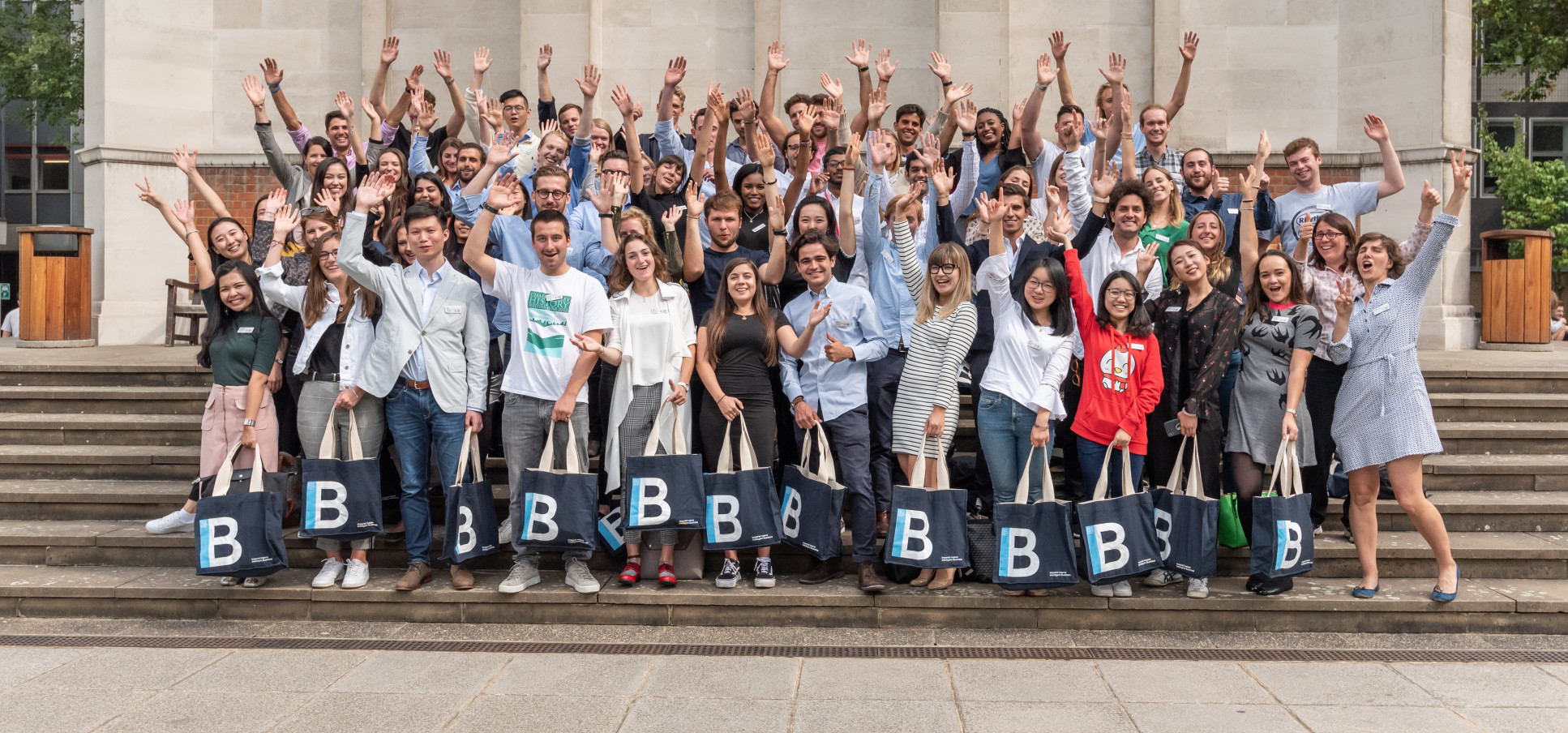 A group of 60 people stand in a group waving their hands and cheering. The front row people people carry matching colourful tote bags reading 'B' for Business  School.