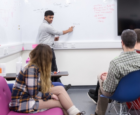 Man writing on a white board while turned to explain notes to a group of sitting students