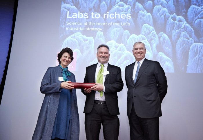 Three people standing on a stage in front of a sign reading 'Labs to Riches'
