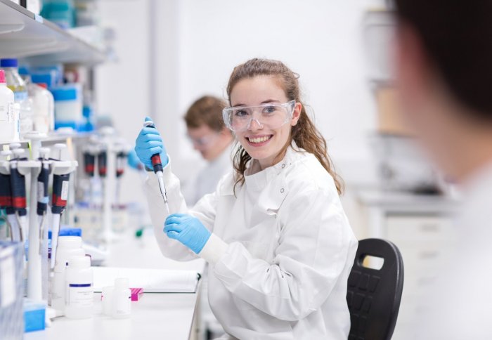 Scientific laboratory icluding a female student with lab coat blue latex gloves and goggles smiling to other