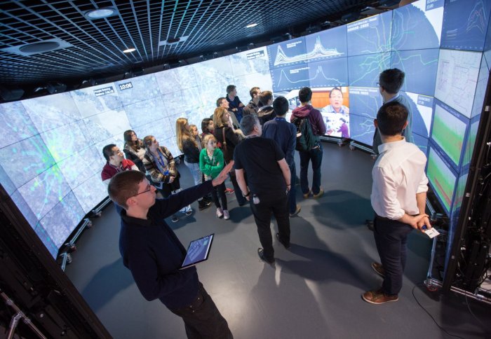 People in a technology room being shown a visual image on a giant screen.