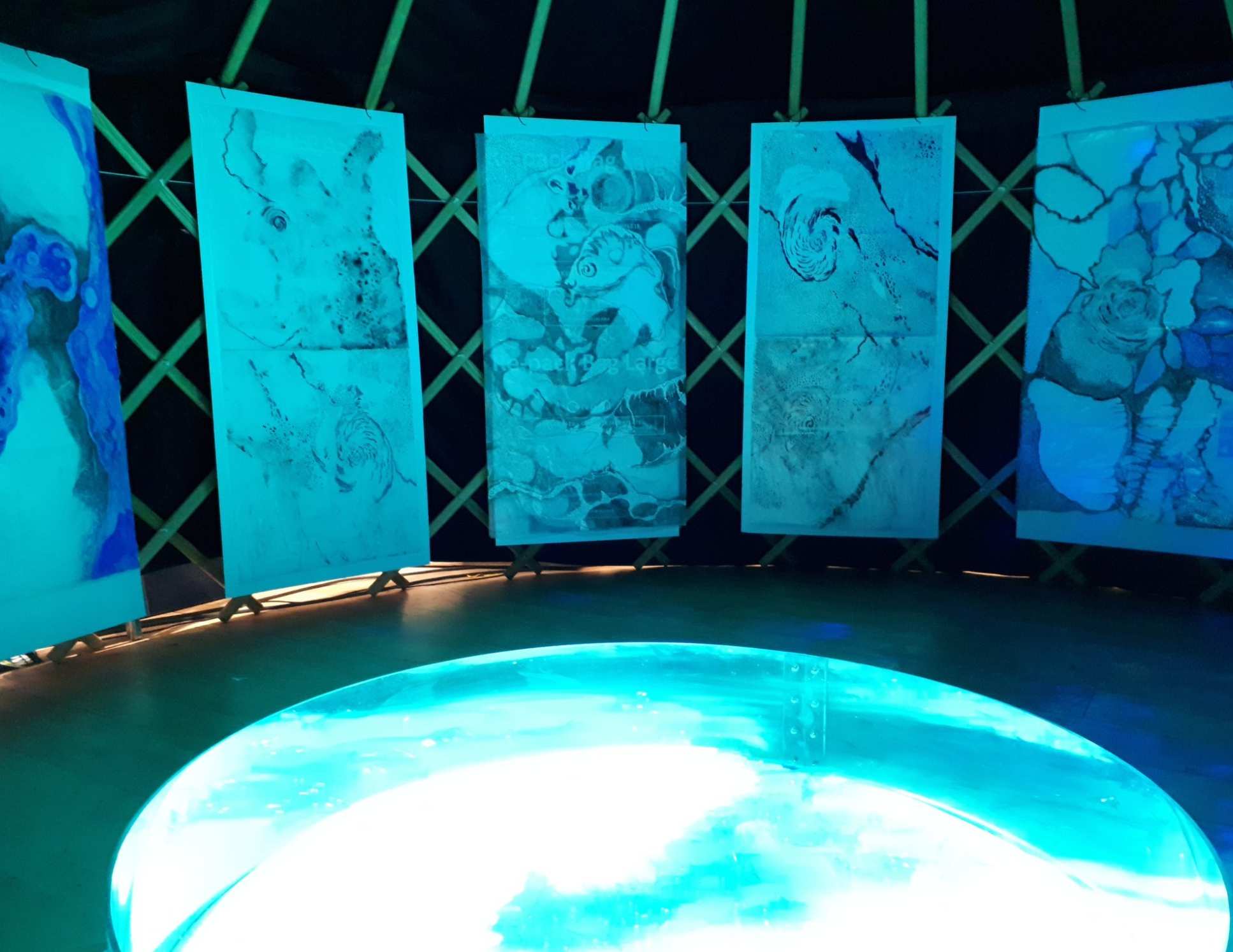 Five abstract watercolour paintings hang above a small pool of water, both are illuminated with blue light