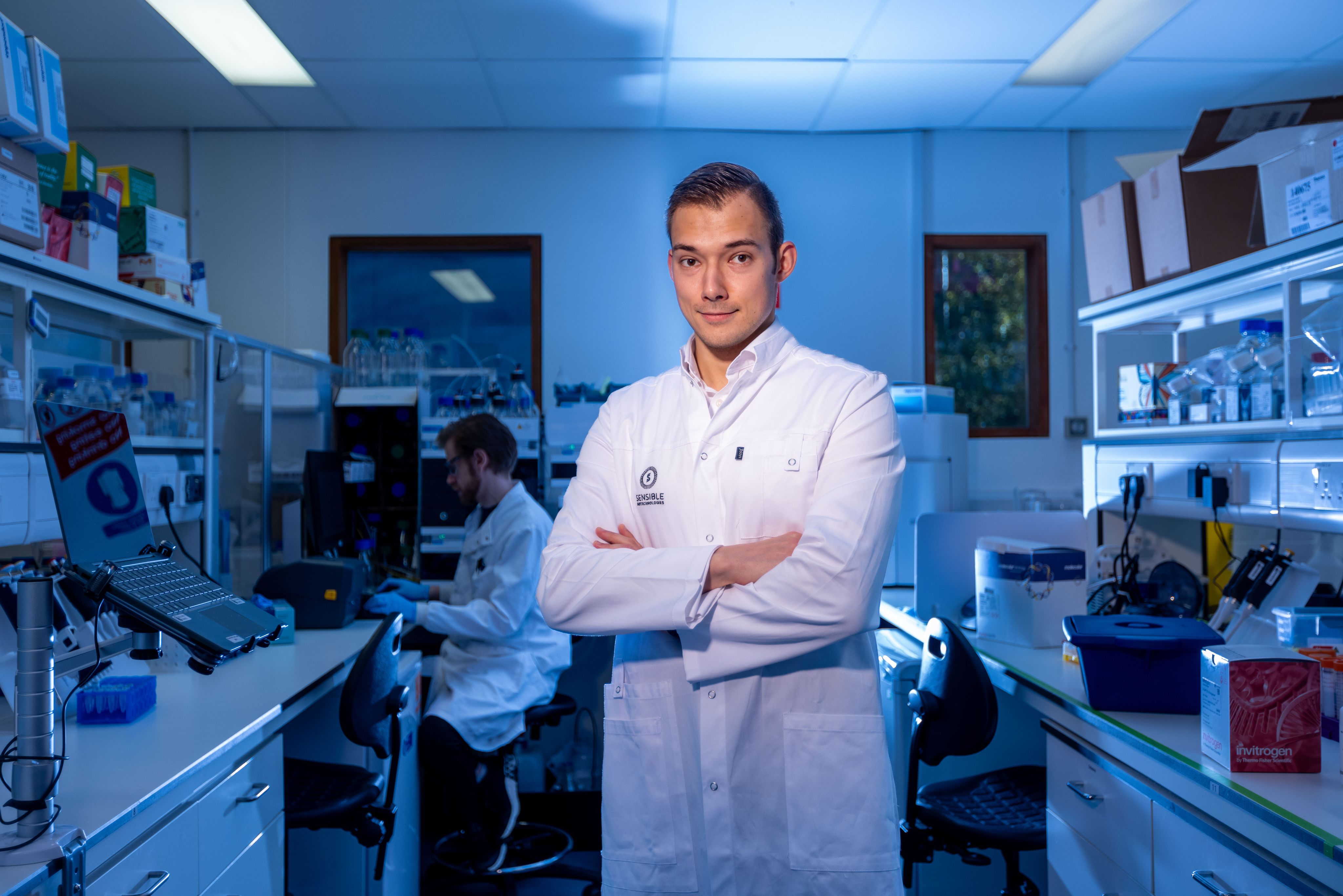 Miroslav standing with his arms crossed in his lab. He is wearing a white lab coat with his company name on it. The lab is illuminated with blue lighting and a colleague works in the background. 