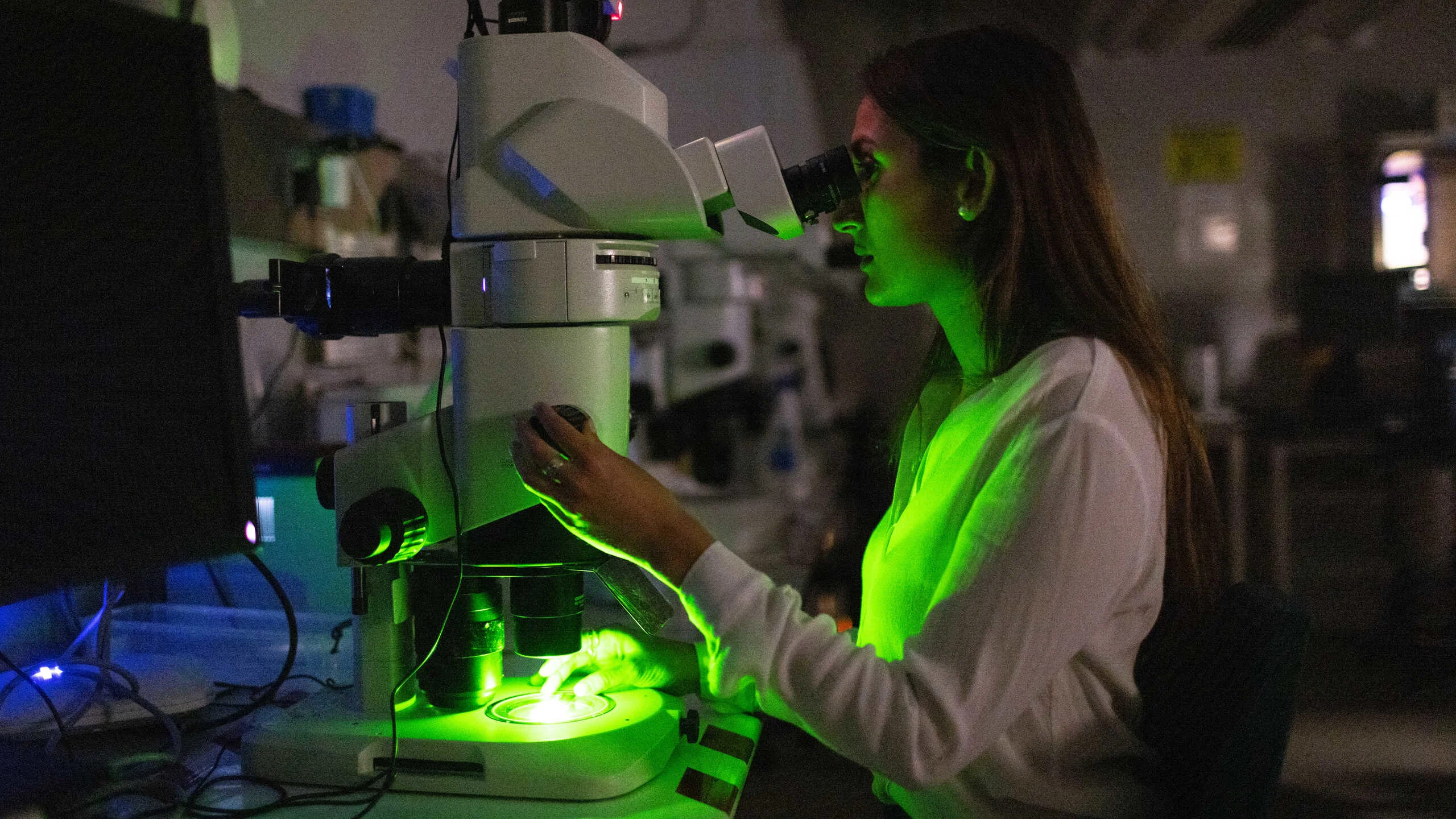 A woman looking down a microscope in a dark room with a green glow
