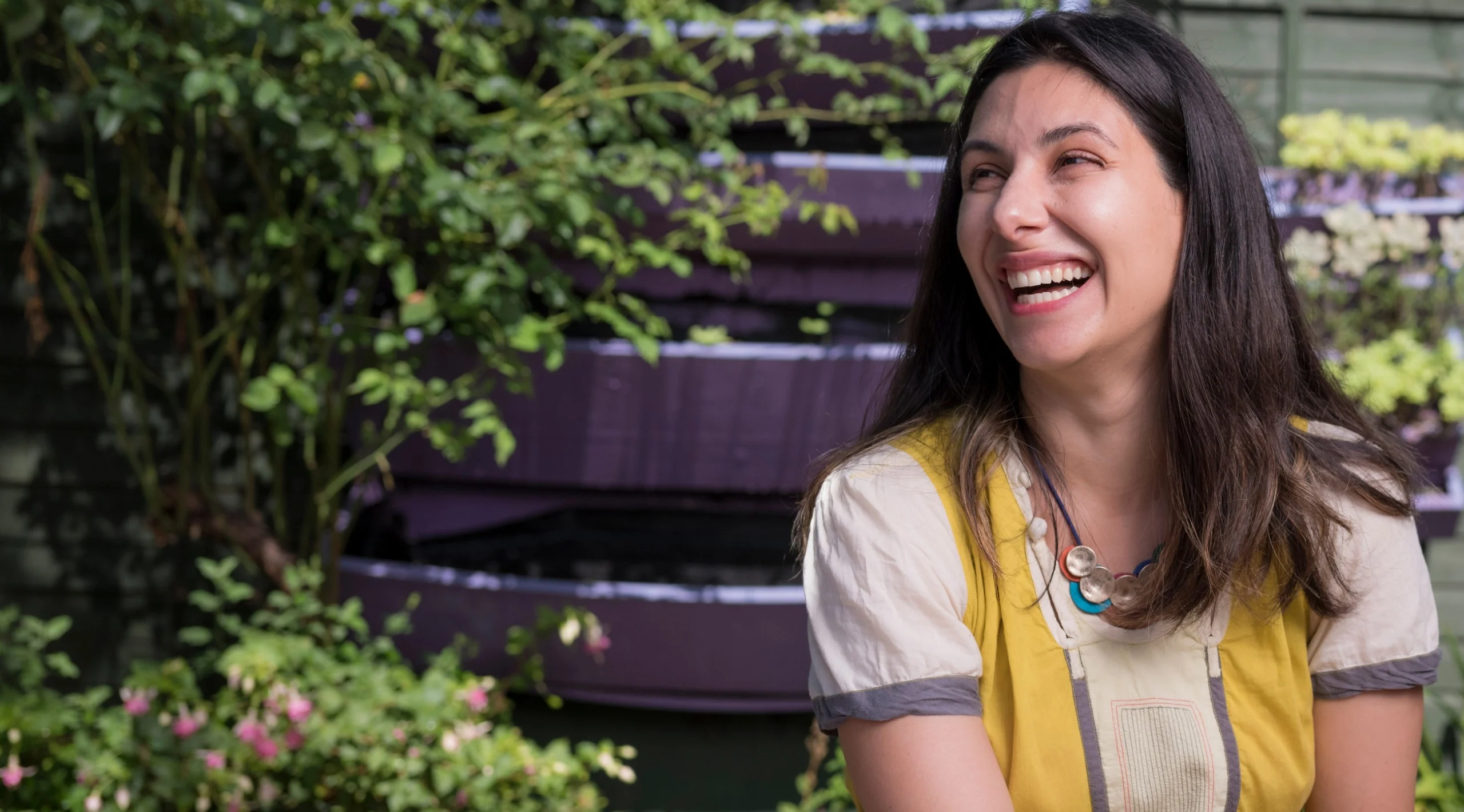 Roya sits on a table in her garden surrounded by flowers. She wears a colourful tunic.