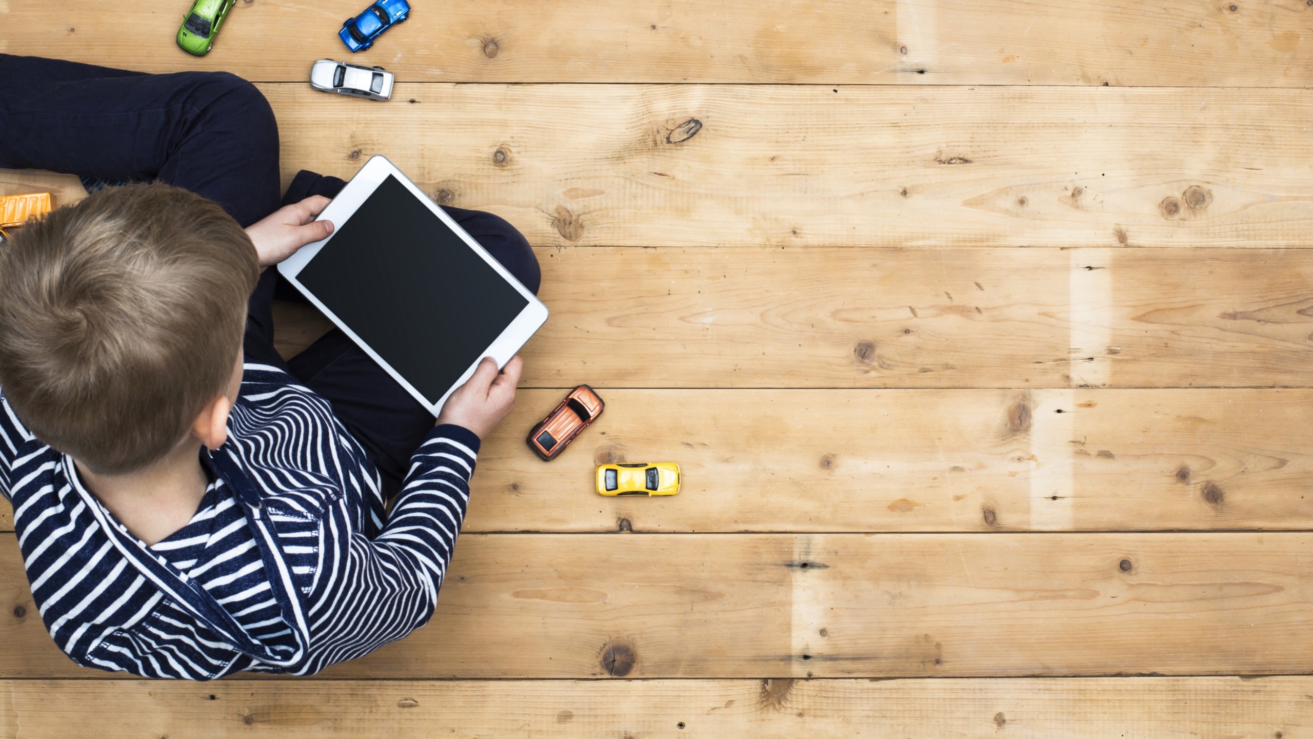 child sits on floor with tablet with small toy cars on around them